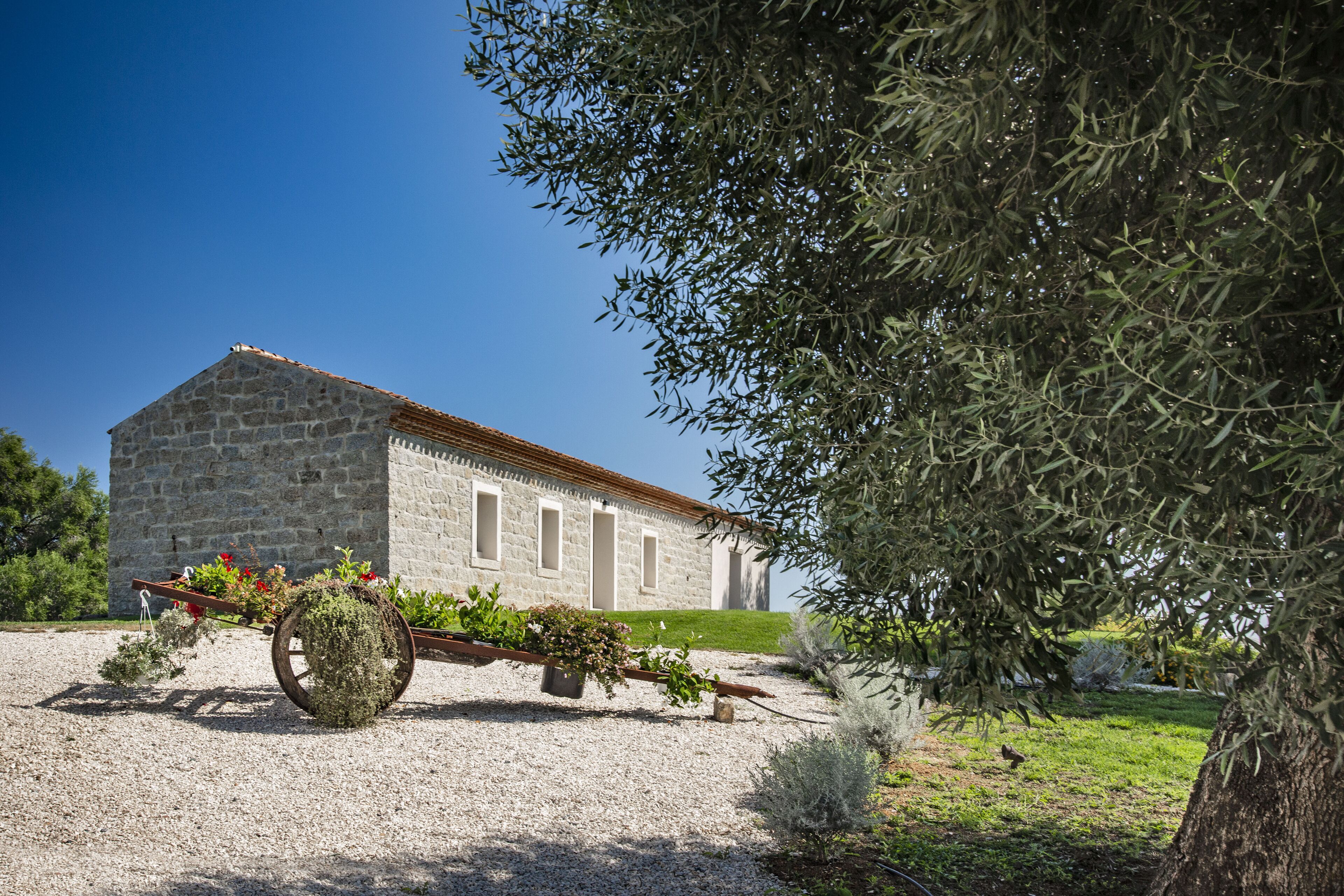 A stone building with a sloped roof, a gravel driveway, and a wheelbarrow with plants.