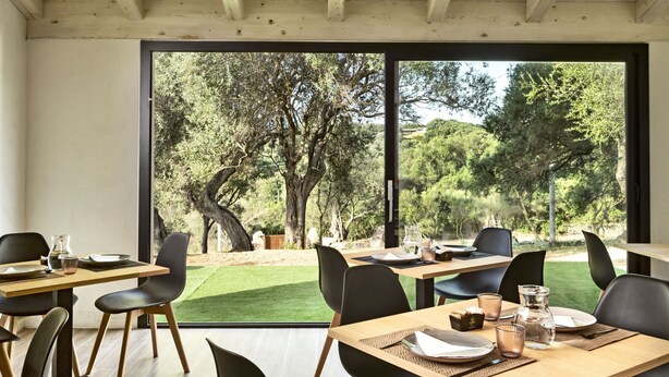 A dining area with tables and chairs, large windows, and a view of trees and grass outside. - Uddastri Retreat (Arzachena)
