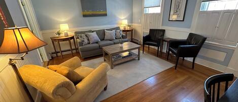 A living room with a sofa, armchairs, a coffee table, and a dining table. There are two lamps, a painting on the wall, and a window with blinds.