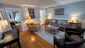 A living room with a sofa, armchairs, a coffee table, and a dining table. There are lamps on side tables and a painting on the wall. - Dive into downtown Newport RI in this private, large, unique townhouse-style 2BR apartment at 205 Spring St! King and Full beds + pull-out sofa full kitchen, 2 baths, outdoor patio, AC, TVs, Wifi. Just steps to Thames Street, shops and wharfs (Newport)
