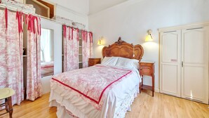 Quarto com cama de madeira, cortinas brancas com estampas florais, mesa de cabeceira de madeira e guarda-roupa.