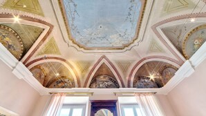 Um salão imponente com afrescos ricamente decorados no teto, um grande candelabro e um detalhado mural floral na parede.