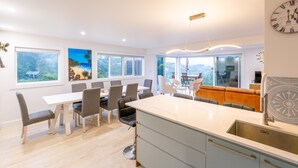 A modern kitchen with a large island, dining area with chairs, and large windows offering views of the outdoors. - Newly renovated 3 bedroom, 2.5 bathroom premium holiday home with stunning views (Waiheke Island)