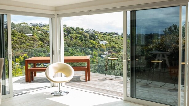 A modern room with a large sliding glass door leading to a balcony with a wooden table and a chair. - Newly renovated 3 bedroom, 2.5 bathroom premium holiday home with stunning views (Waiheke Island)