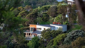 A modern house surrounded by dense greenery, with a prominent balcony and a visible satellite dish. - Newly renovated 3 bedroom, 2.5 bathroom premium holiday home with stunning views (Waiheke Island)