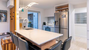 A modern kitchen with a central island, stainless steel appliances, and a dining area with a wooden table and chairs. - Newly renovated 3 bedroom, 2.5 bathroom premium holiday home with stunning views (Waiheke Island)