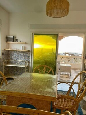 Uma área de refeições com uma mesa e cadeiras, uma prateleira pequena com um aparelho, e vista através de uma porta de vidro.