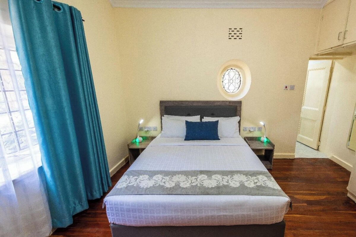 A neatly made bed with a patterned bedspread, two bedside tables with lamps, a window with a decorative grille, and a door to the right.