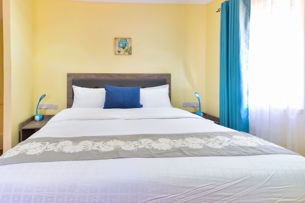 A neatly made bed with white and blue bedding, a decorative pillow, and a floral artwork on the wall.