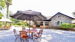 Une terrasse extérieure avec une table ronde et des chaises, un grand parasol, et un bâtiment en pierre avec des fenêtres en arrière-plan.