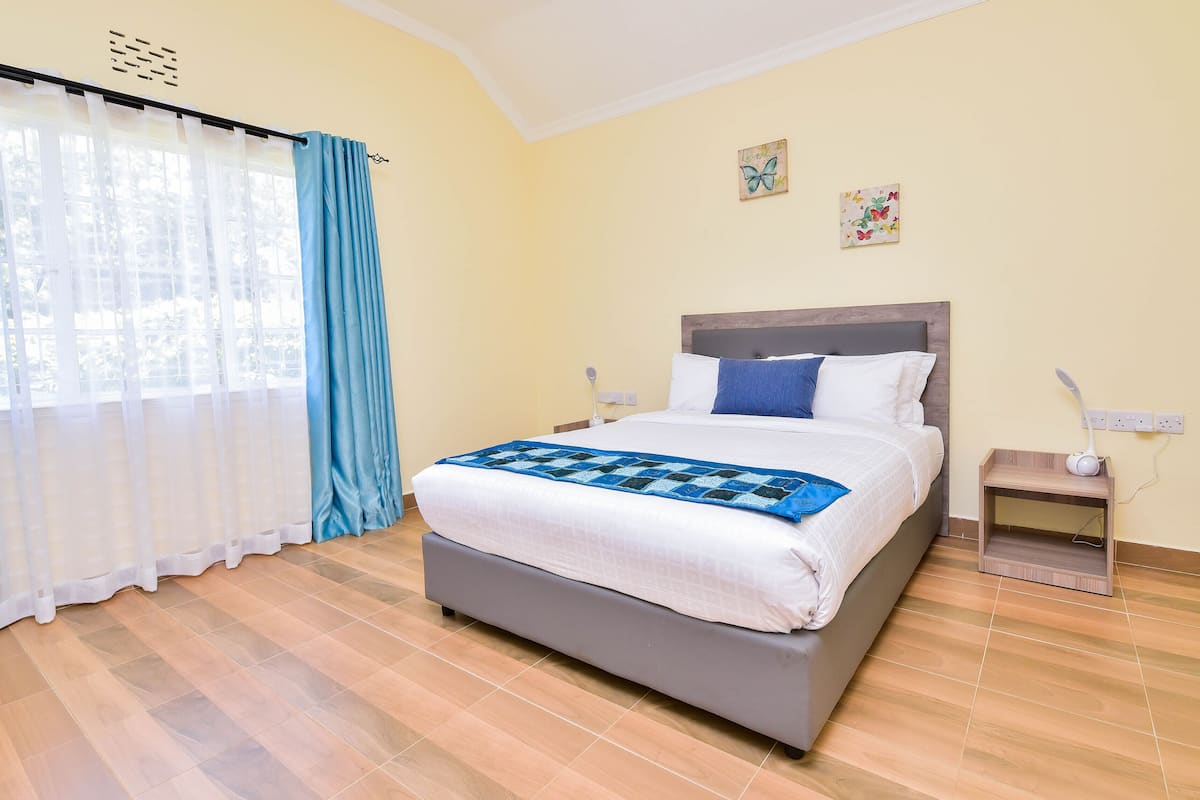 A bedroom with a bed, curtains, a bedside table, a lamp, and a wall with two small pictures.
