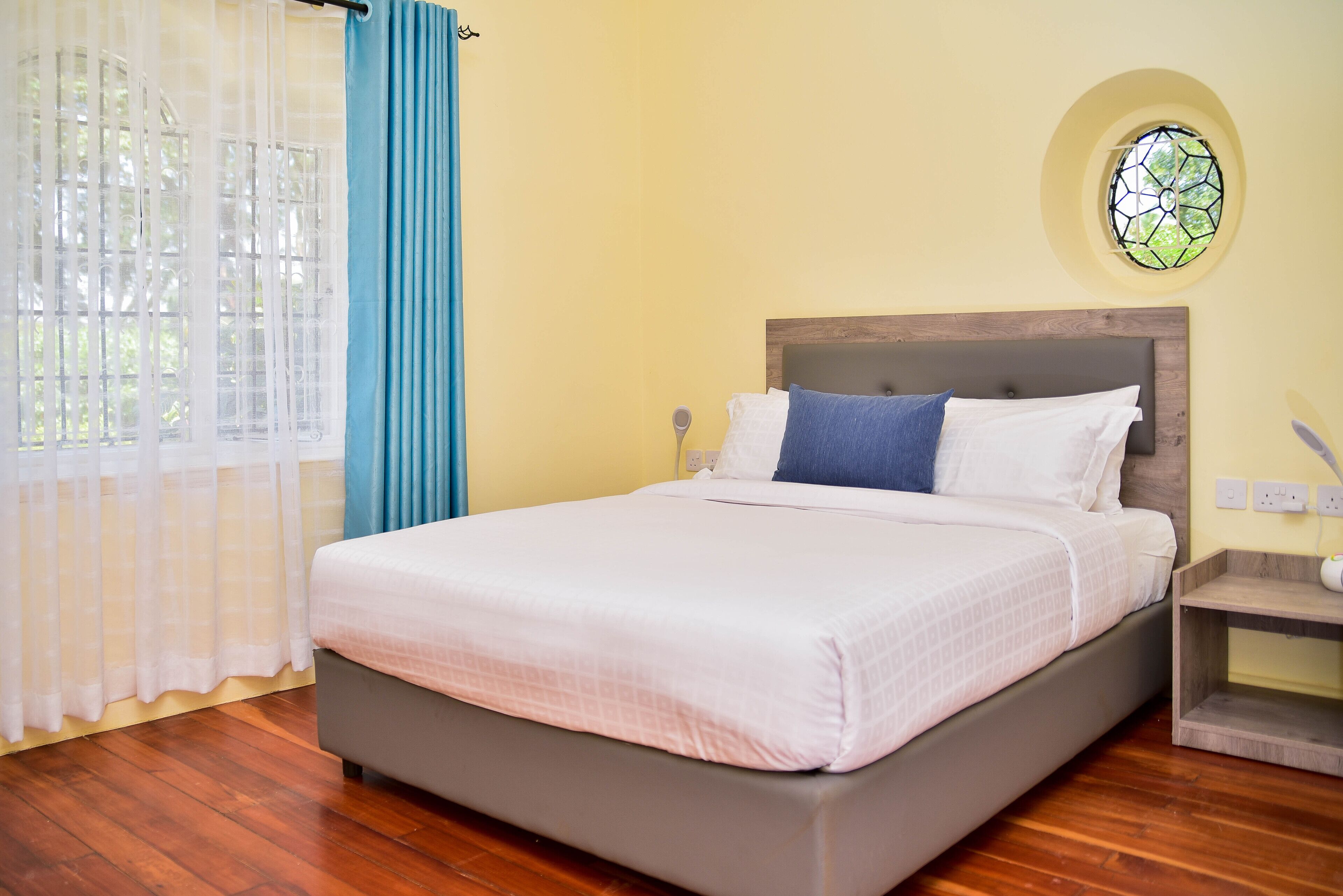 A bedroom with a bed, wooden floor, a nightstand, a wall-mounted light, and a window with a curtain.