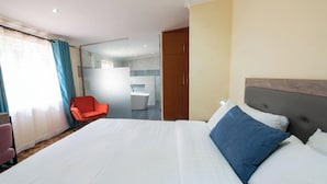 Une chambre d’hôtel avec un grand lit, une chaise, une salle de bain visible à travers une paroi en verre et une armoire.