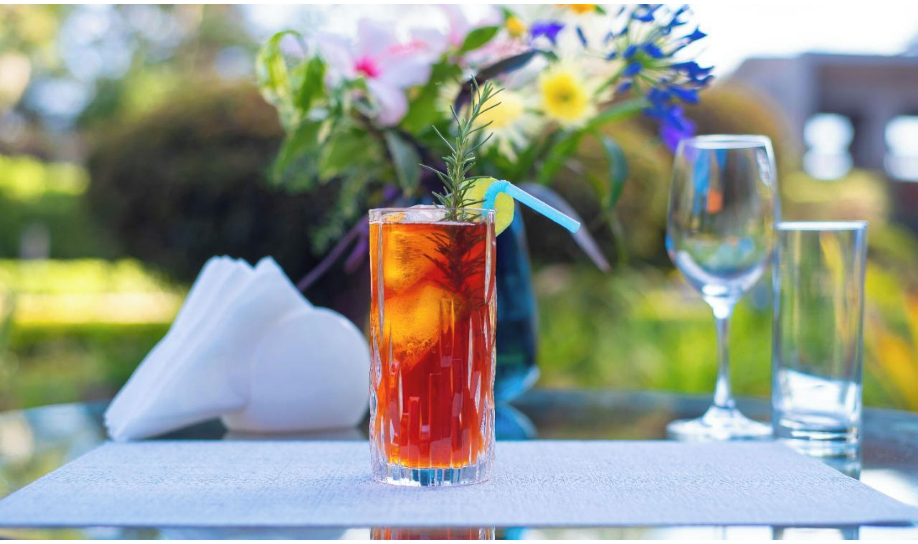 A tall glass with a red beverage, a clear glass, and a napkin on a table with a floral arrangement in the background.