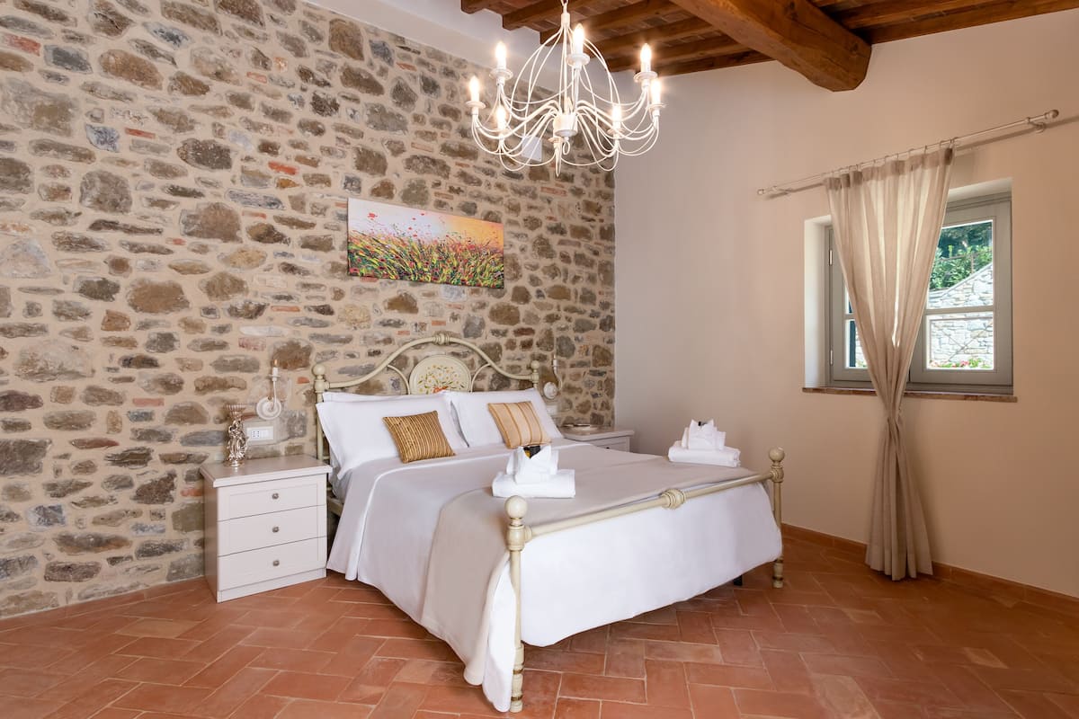 A bedroom with a stone wall, a chandelier, a bed with white linens, a nightstand, and a window with curtains.
