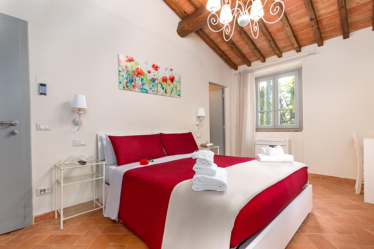 A hotel room with a bed, a window, a door, a nightstand, and a painting on the wall.
