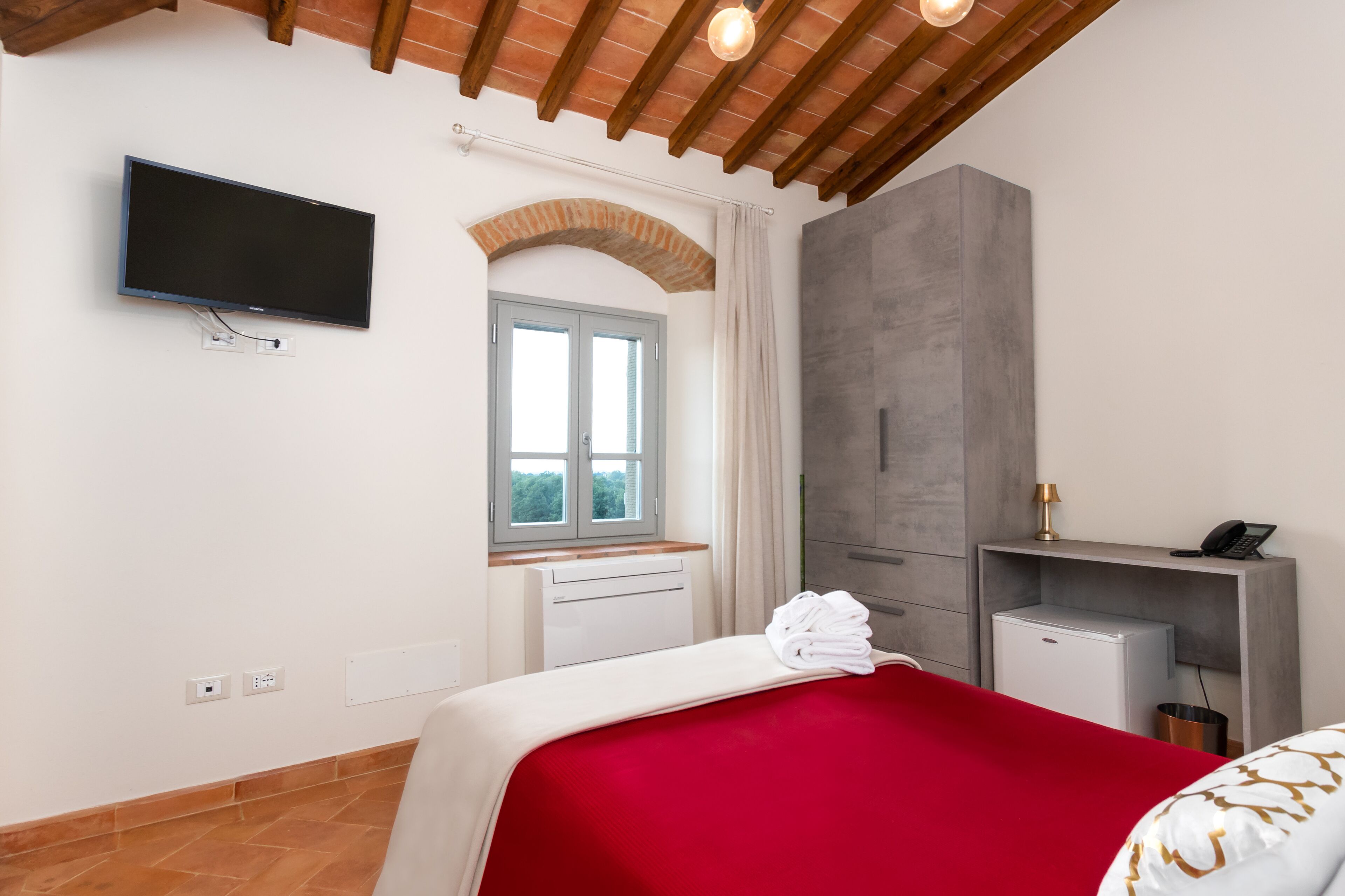 A hotel room with a bed, a TV mounted on the wall, a window with curtains, a small desk with a lamp, and a minibar.