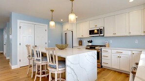 A modern kitchen with white cabinetry, a marble island, wooden chairs, and pendant lighting. - On Par for the Beach (Old Orchard Beach)