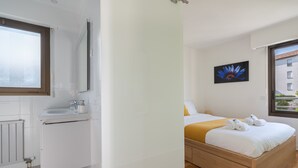 A modern hotel room with a wooden bed, white walls, a large window, and a bathroom with a white sink and mirror. - LE PARC Apartment for 5 By Sunset (Antibes)
