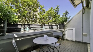 Eine kleine Terrasse im Freien mit einem runden Tisch und Stühlen, einem Blumentopf und Blick auf ein Gebäude mit weißer Tür und Fenster.