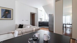 Ein moderner Wohnbereich mit Sofa, Couchtisch mit zwei Tassen, Küchenzeile mit Schränken und Kühlschrank sowie einem gerahmten Bild an der Wand.