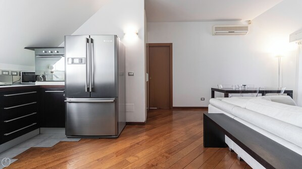 A modern kitchen with a stainless steel refrigerator, a microwave, and a coffee maker. There is a wooden floor, a bed with white bedding, and a desk with a chair.