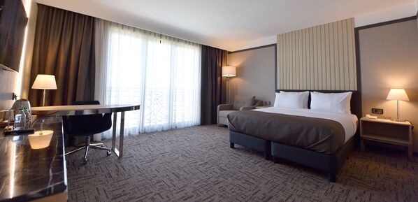 A hotel room with a bed, bedside tables, a desk with a chair, and a large window with curtains.
