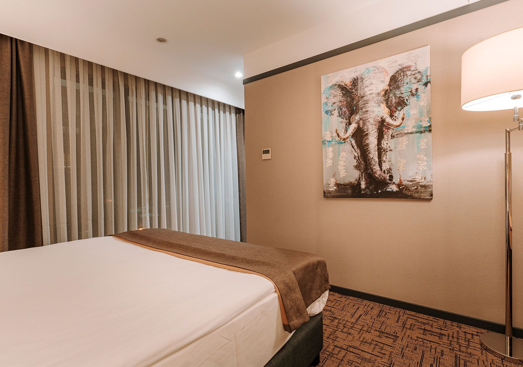 Una camera d'albergo con un letto, un'ampia finestra con tende, un quadro appeso al muro e una lampada da terra.