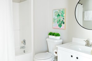 Bathroom features a white bathtub, toilet, and sink, with a framed painting of tropical leaves on the wall.