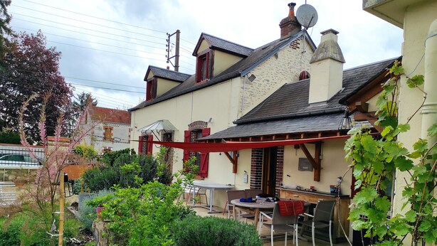 A house with a patio area, a dining table, and chairs. There is a garden with various plants and a small tree. - A 1h de Paris, et 1h des Châteaux de la Loire, (CHALETTE SUR LOING)