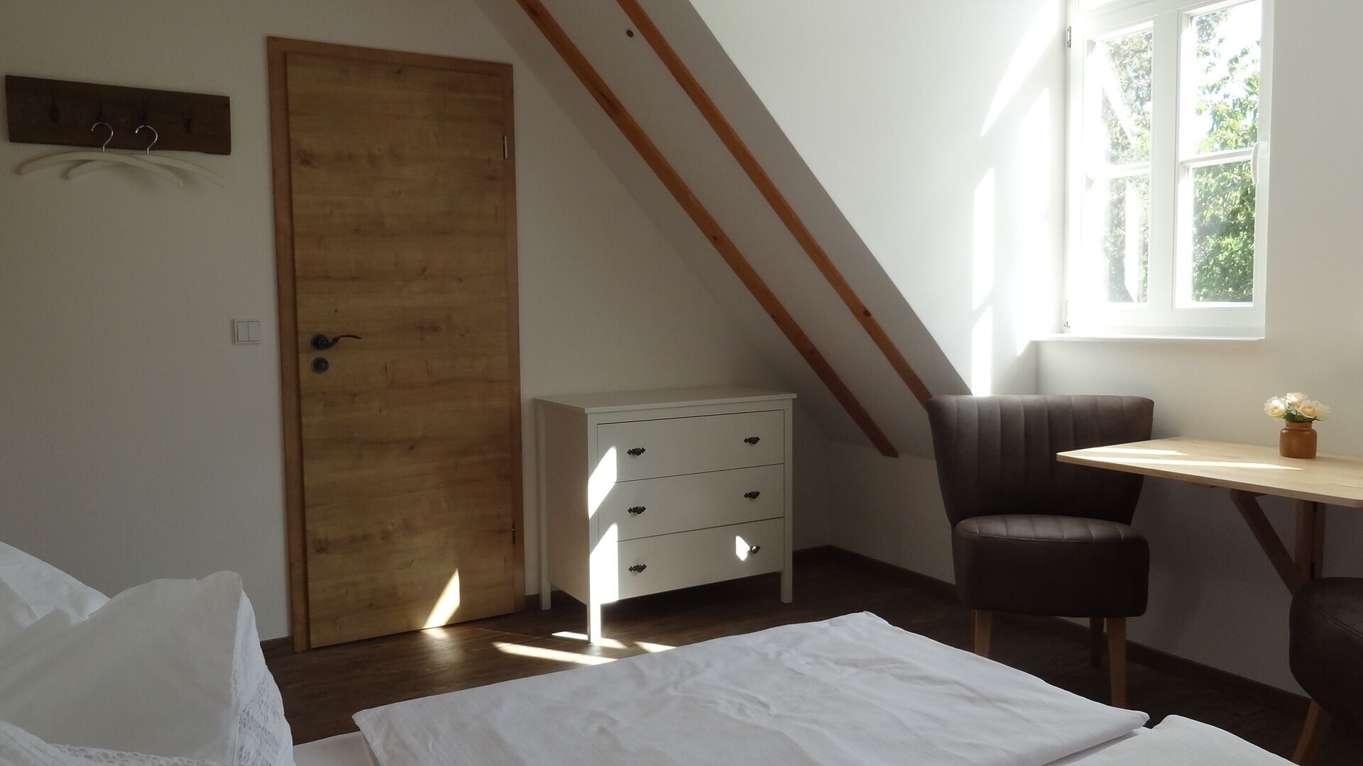 Ein Schlafzimmer mit Holztür, weißem Schminktisch, Schreibtisch, Stuhl und Fenster mit Blick auf Bäume.