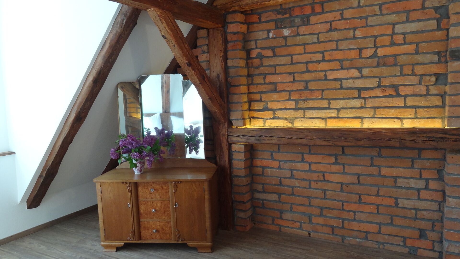 Un mueble de madera con cajones y un florero con flores moradas, ubicado frente a una pared de ladrillo con estructura de vigas de madera.