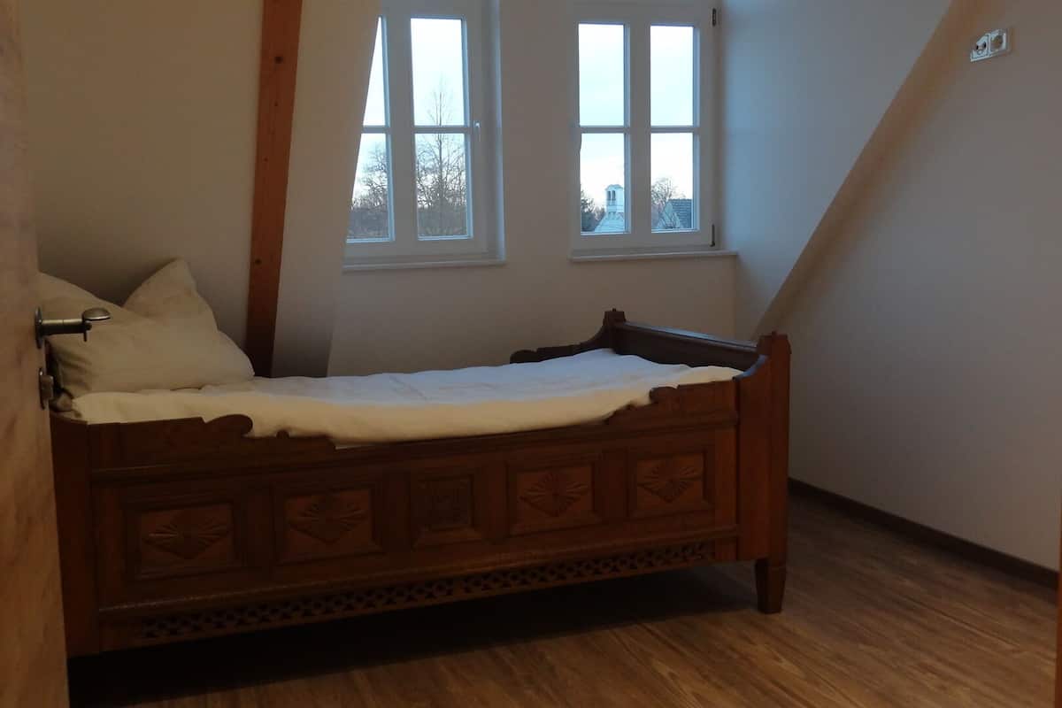 Una habitación pequeña con cama individual, techo inclinado y dos ventanas.
