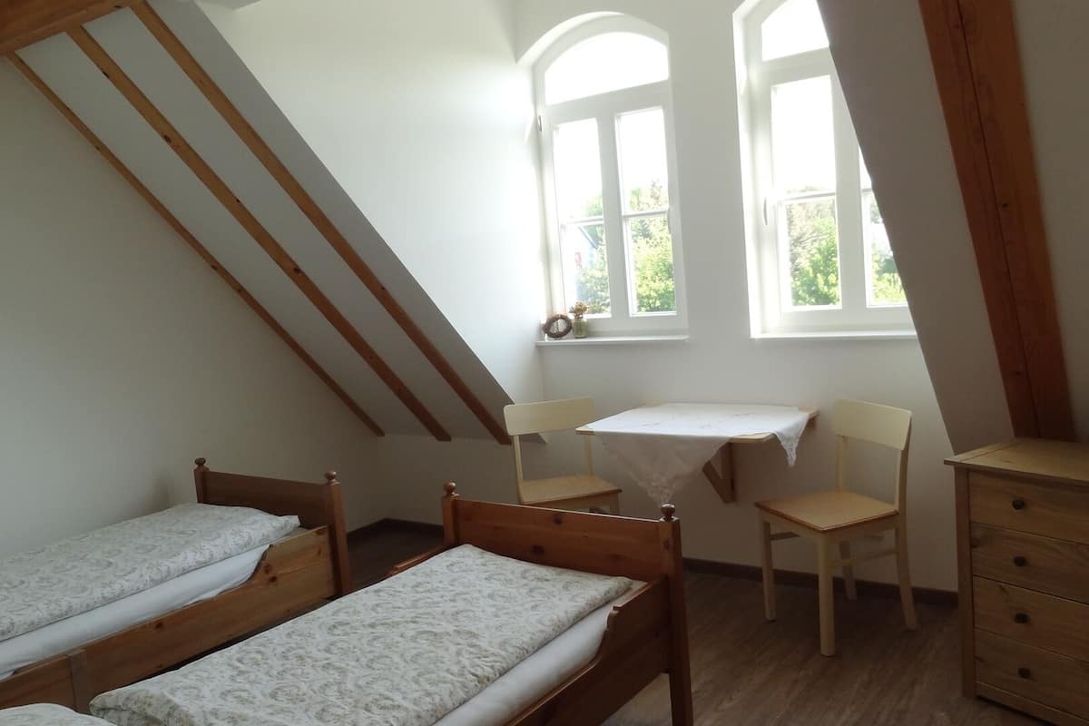 Un dormitorio con dos camas de madera, una mesita, dos sillas, un aparador y una ventana con vistas a zonas verdes.