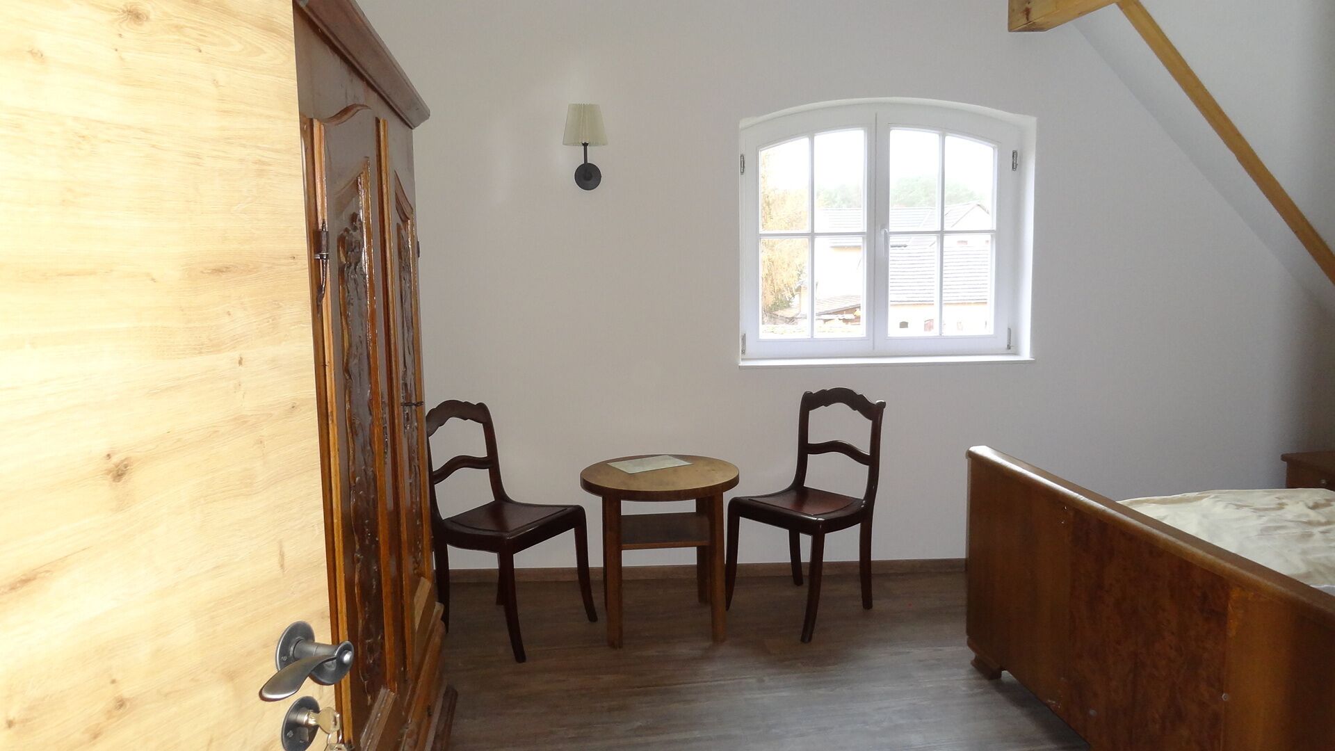 A small, cozy room with a wooden door, two chairs, a small round table, a bed, and a window with a view of the outside.