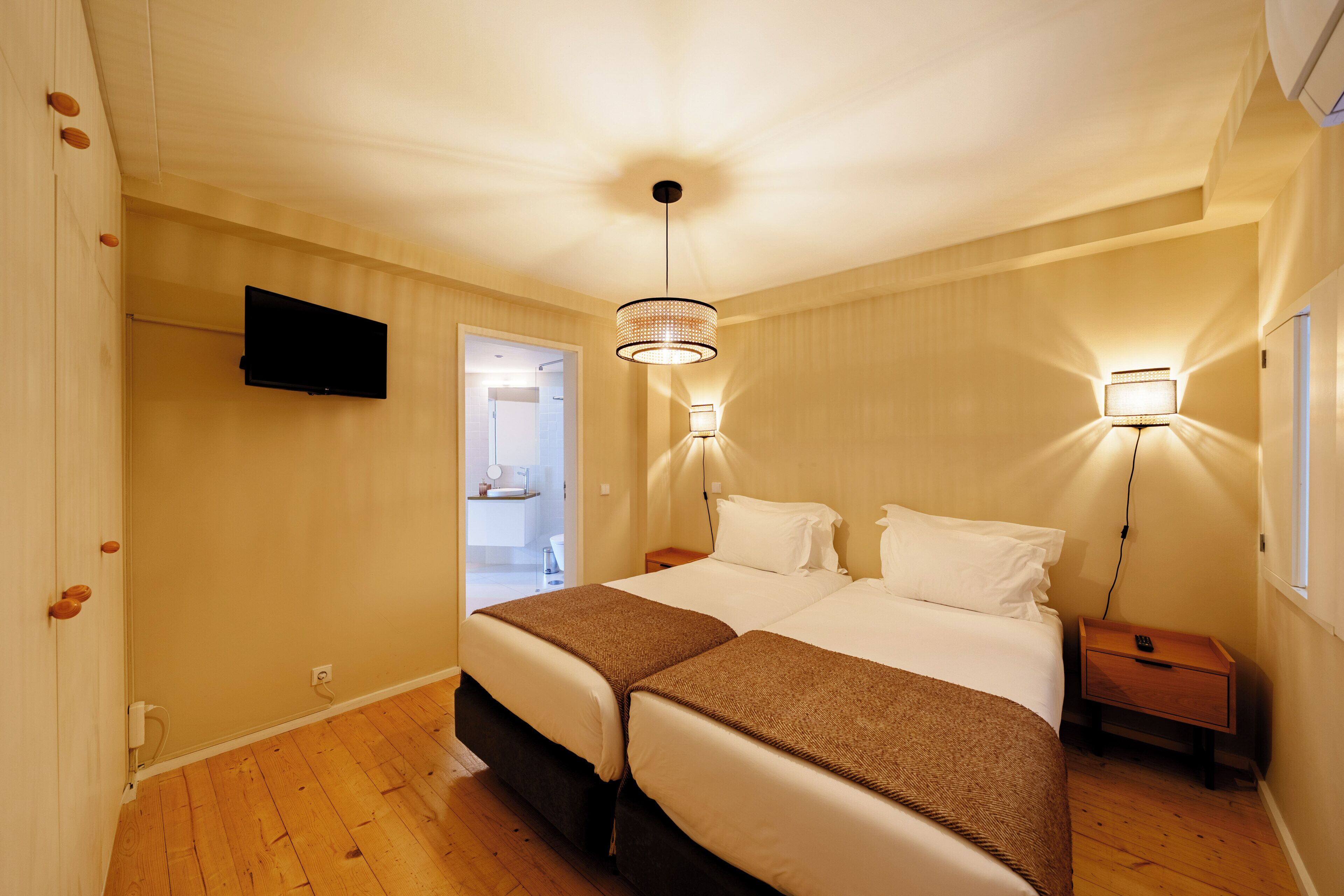 Quarto de hotel com duas camas, televisão na parede, abajures e chão de madeira.