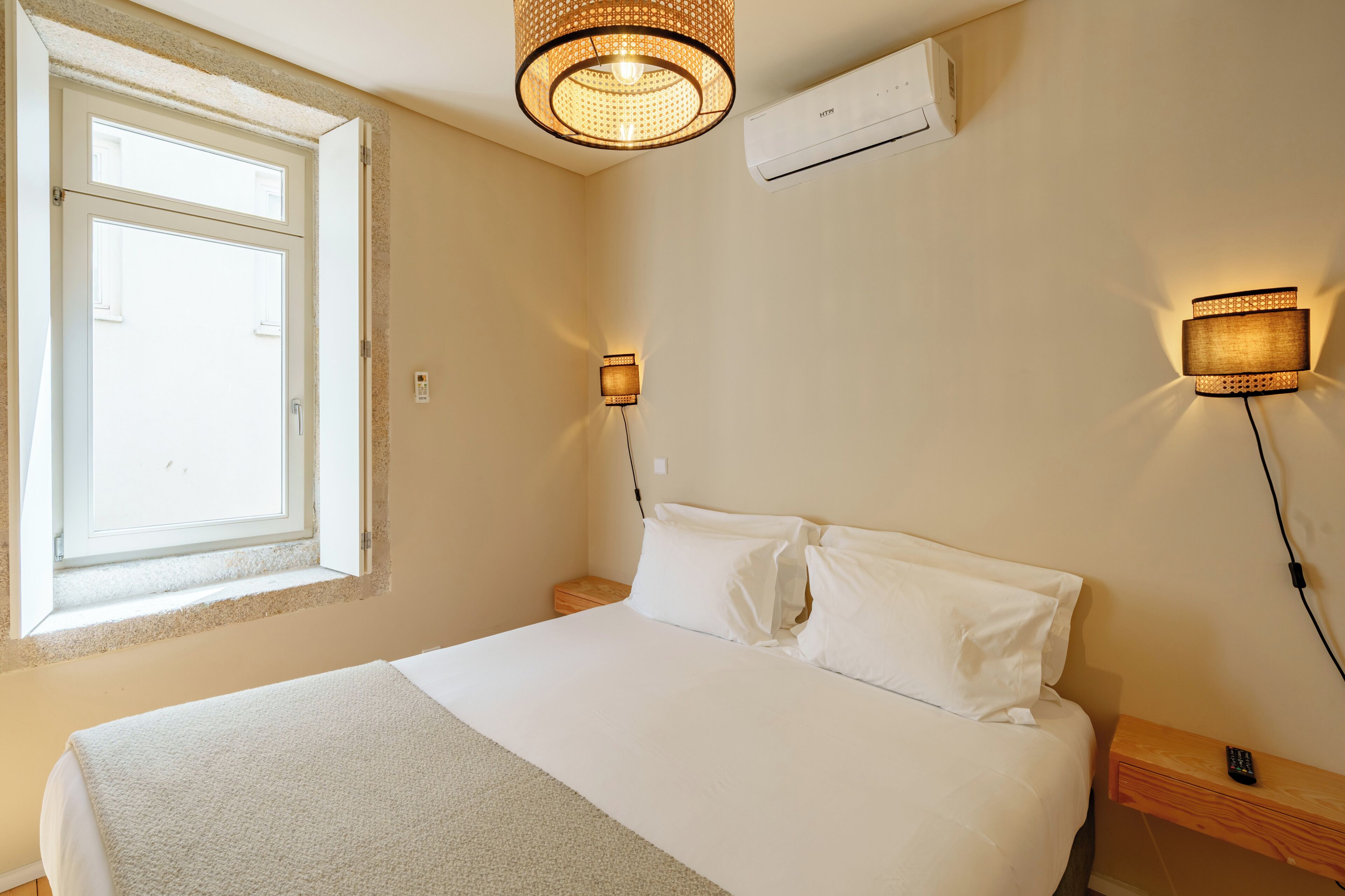 A bedroom with a bed, a window, a wall-mounted air conditioner, and a wall-mounted lamp.