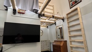 A compact room with a bunk bed, a flat-screen TV, a vanity with a mirror, a wooden dresser, and a ladder.