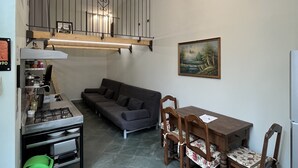 A compact kitchen with a gas stove, a dining table with chairs, a sofa, and a hanging light fixture.