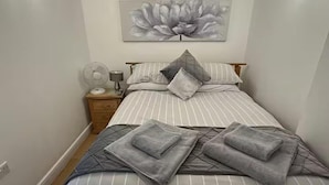 A neatly made bed with striped bedding and pillows, a bedside table with a lamp and a fan, and a large floral artwork on the wall. - Hertford - Sleeps 2 - 92 Walking Score - Garden (Hertford)