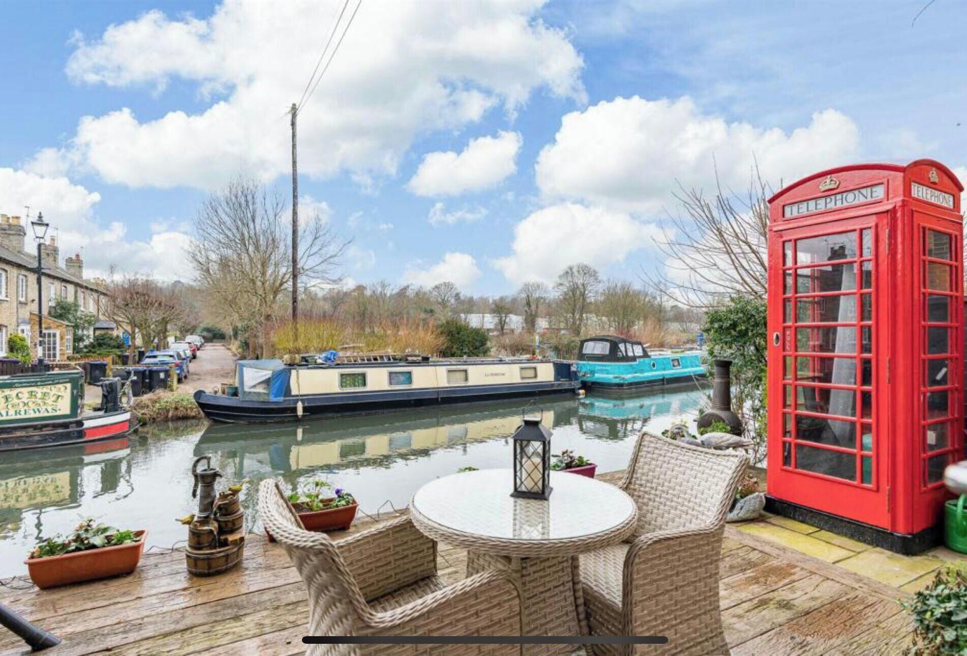 Un cadre pittoresque au bord de la rivière, avec une cabine téléphonique rouge, des meubles en osier et des bateaux sur l’eau.