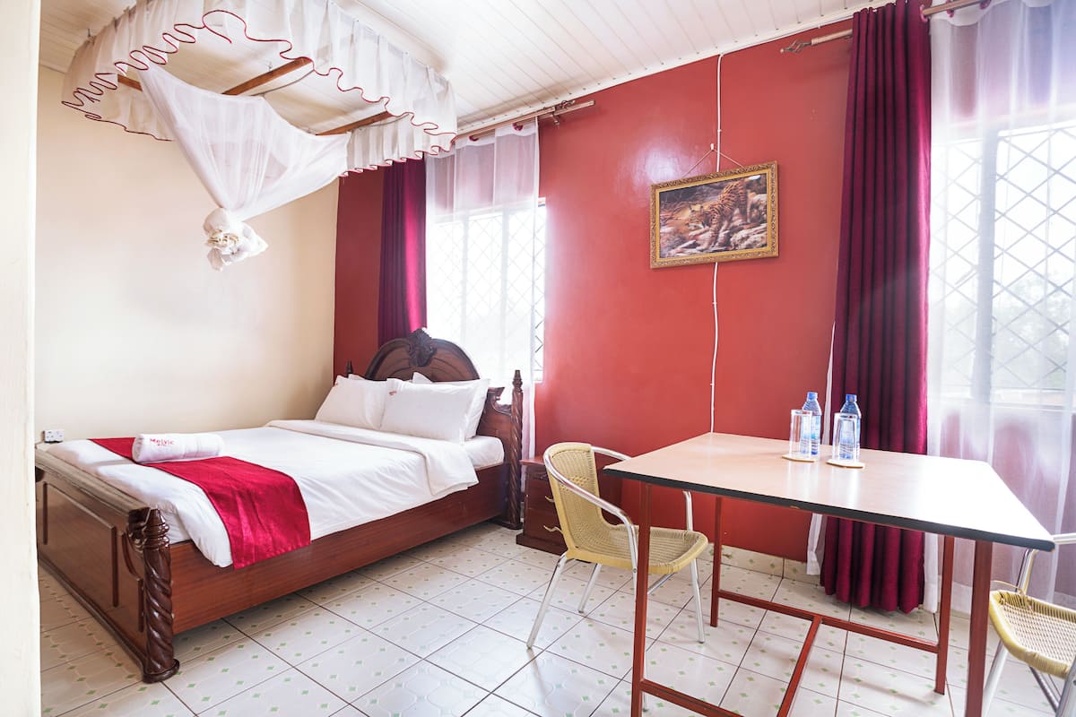 Ein Zimmer mit einem Bett, einem Tisch, Stühlen, einem fenster mit Vorhängen und einem Gemälde an der Wand.