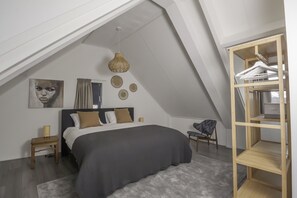Une chambre avec un plafond en pente, un lit recouvert d’une couvre-lit gris foncé, une table de chevet en bois, une chaise et une étagère en bois où sont pliés des vêtements.