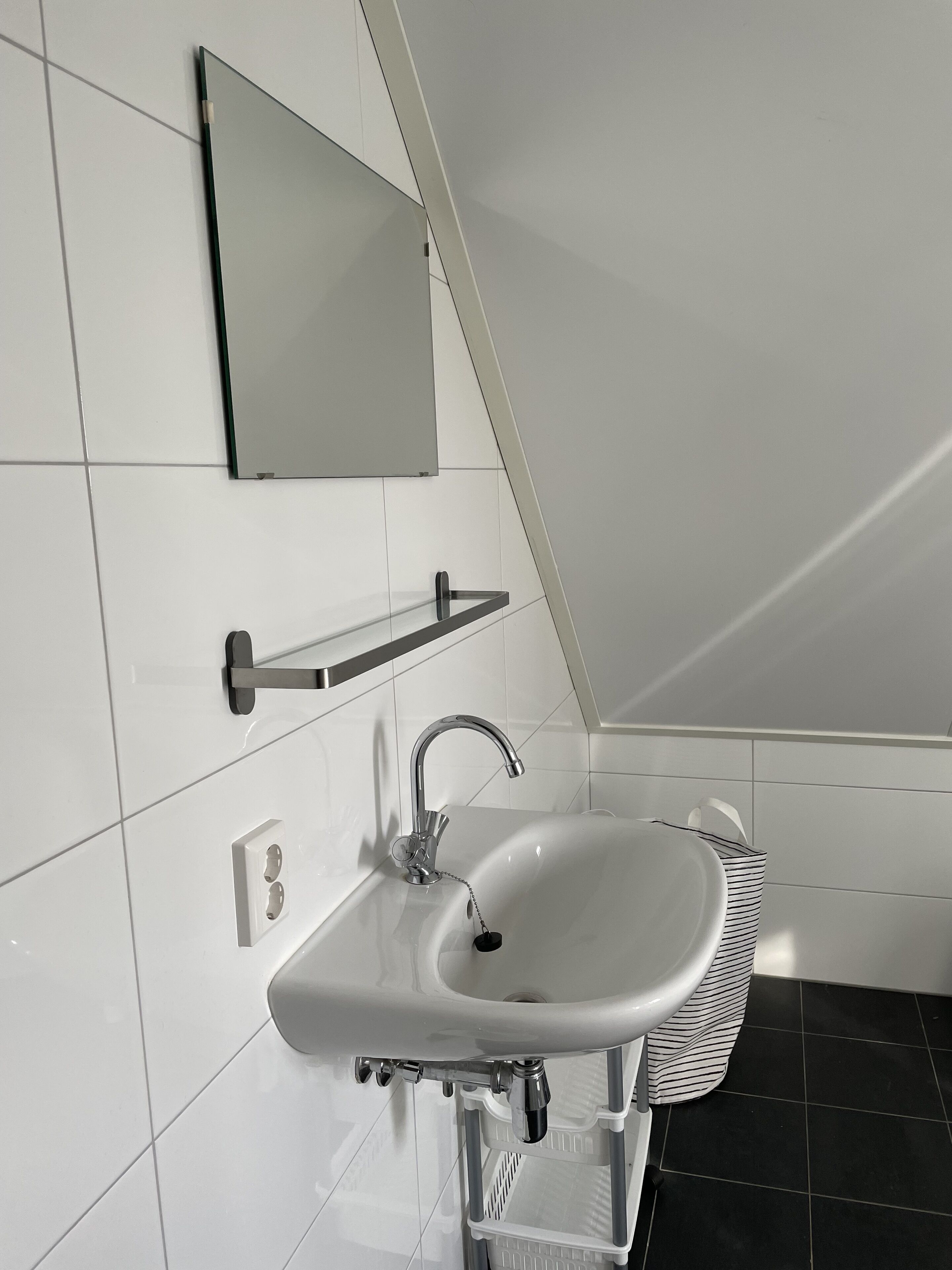 A modern bathroom with a white sink, chrome faucet, and a mirror above it. There is a towel rack and a basket on the side.