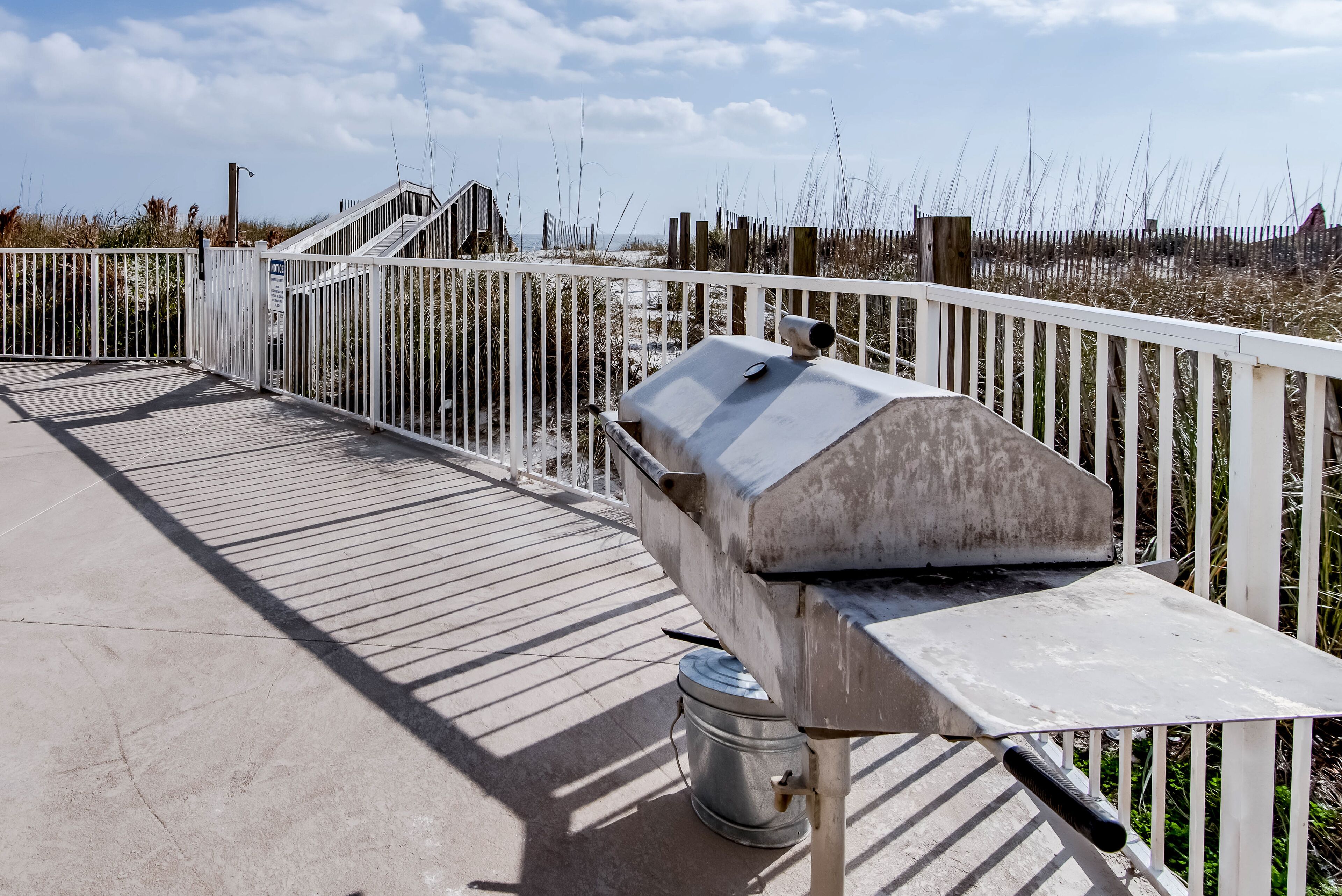 Una parrilla metálica sobre una terraza de concreto con una valla blanca y una playa al fondo.
