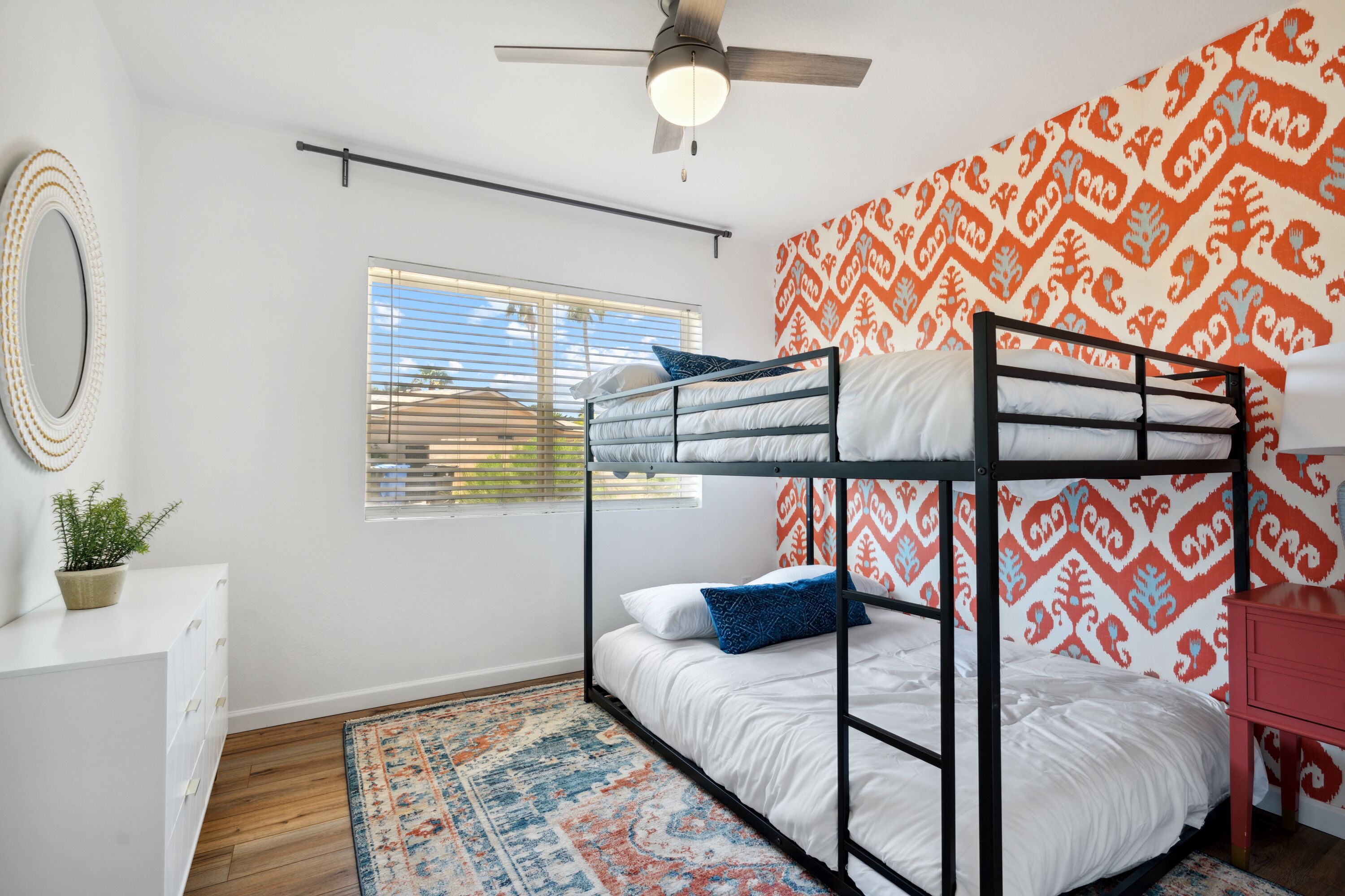 A bunk bed room with a ceiling fan, a window with blinds, a patterned wall, a small desk, and a potted plant.