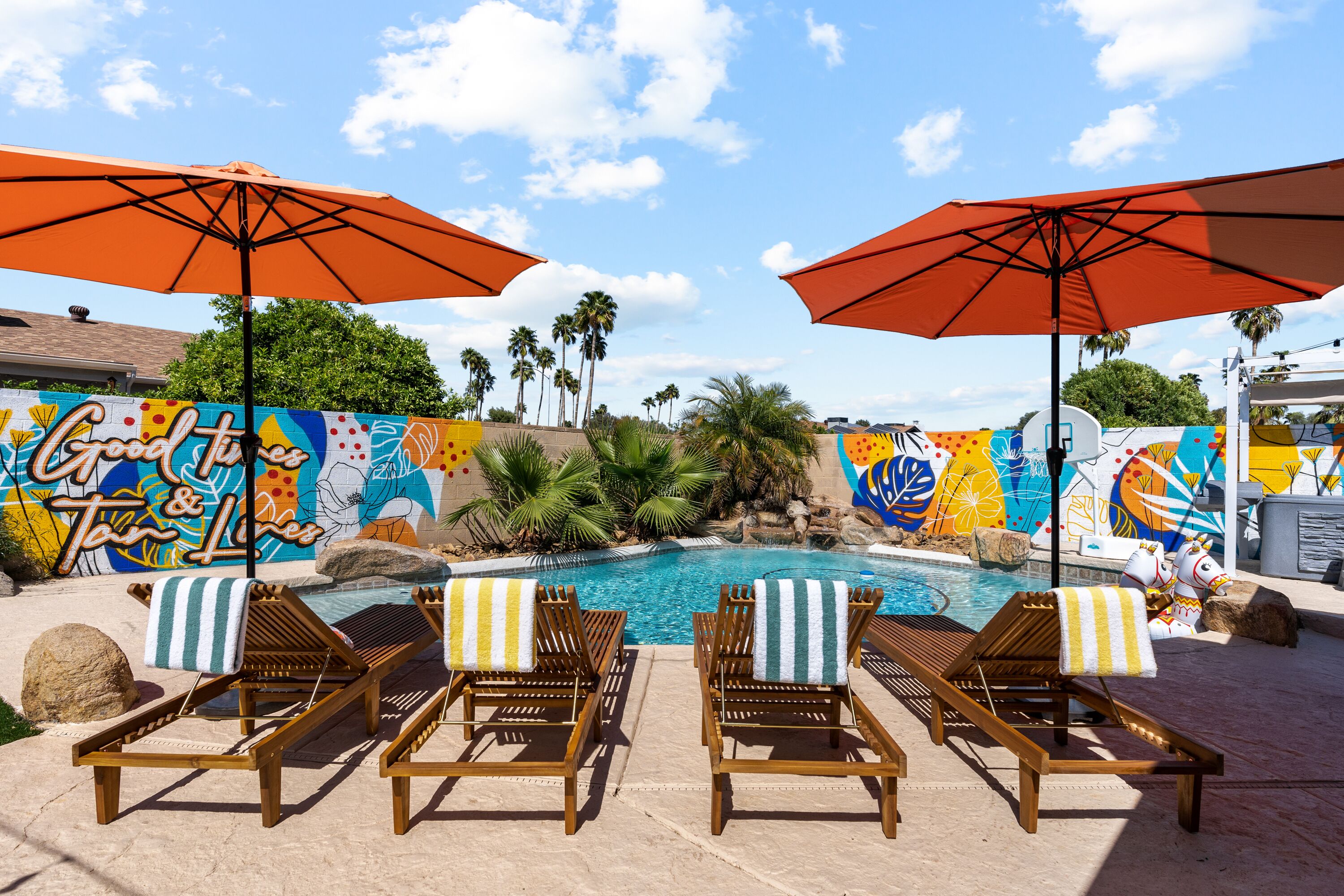 A poolside area with wooden lounge chairs, orange umbrellas, and a colorful mural.