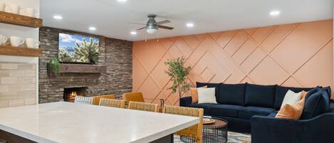 Una cocina moderna con una isla central, un comedor con sillas de madera, un sofá azul oscuro y una chimenea.