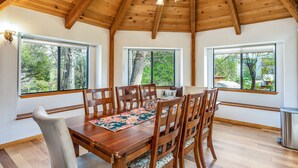 Un comedor con mesa y sillas de madera, ventanas amplias con vistas a árboles y techo de madera.