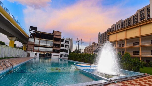 A swimming pool with a fountain, surrounded by buildings and a bridge. - Aristro Club (Alipur)
